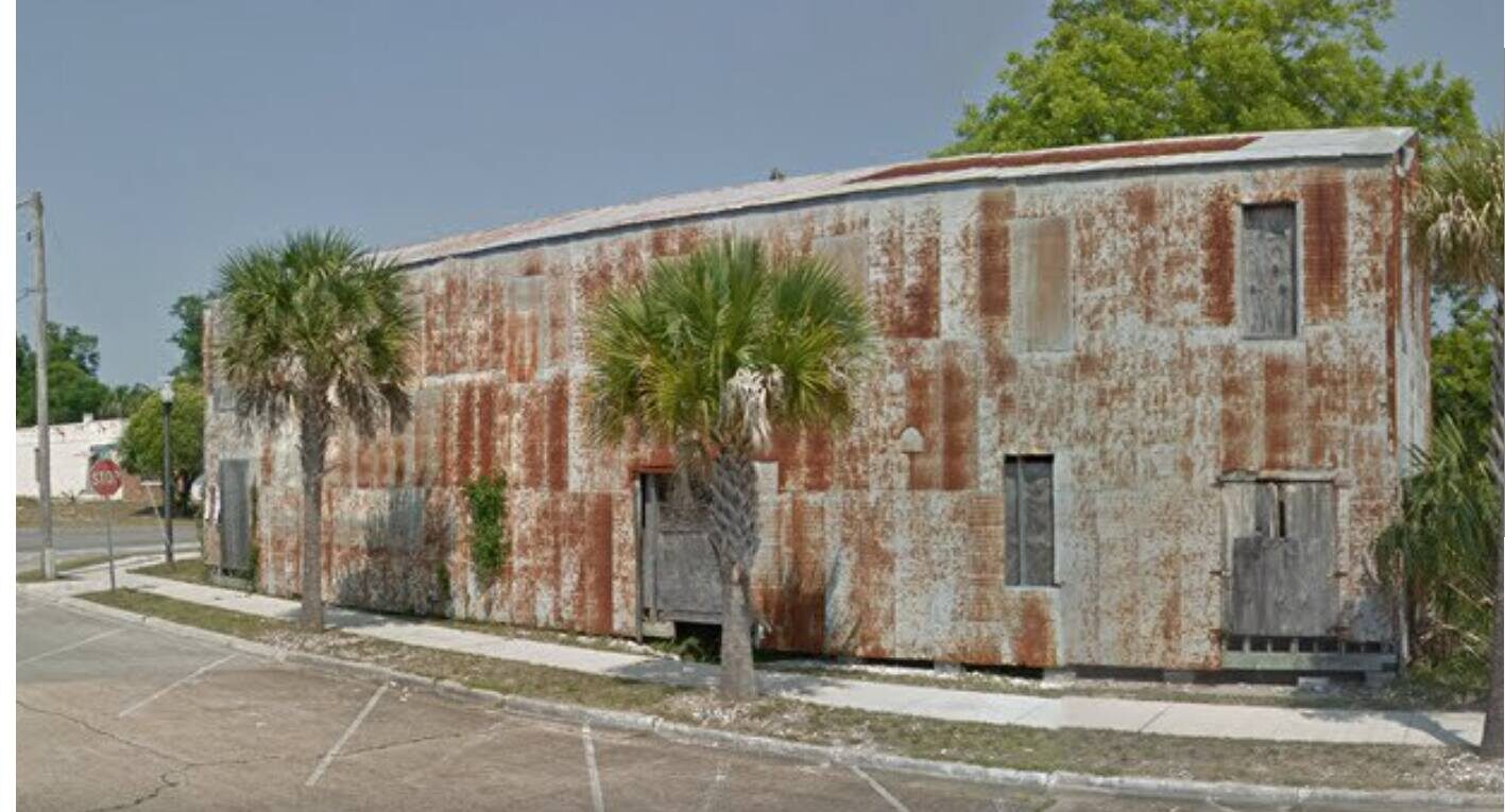 A weathered, rust-colored building with a flat roof and palm trees in front.